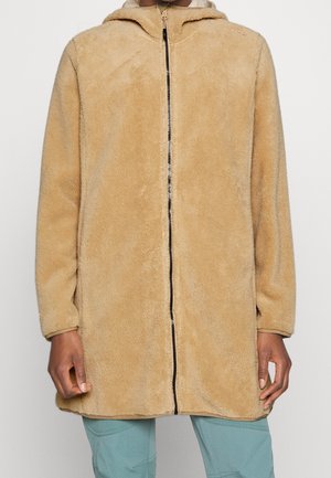 Osoba ubrana w beżową, rozpinaną bluzę polarową z kapturem i długimi rękawami oraz jasnoniebieskie spodnie, stojąca na jednolitym tle.