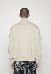Sudadera beige con cuello alto, puños y dobladillo acanalados. Tejido suave, corte holgado, llevada sobre jeans negros desgastados. Presenta un detalle mínimo.