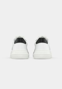 Un paio di sneaker bianche basse con inserto nero sul tallone, con la scritta "Calvin Klein Jeans" in rilievo su suole in gomma spessa, mostrate da dietro su sfondo bianco.