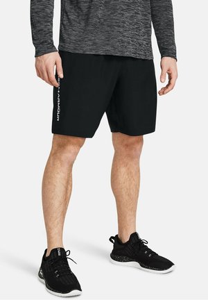 Mann trägt schwarze Sportshorts mit weißem "Under Armour"-Logo an der Seite, schwarze Laufschuhe und ein graues, strukturiertes Langarmshirt.
