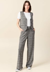 Grahamusterweste mit fünf Knöpfen über einem weißen T-Shirt, kombiniert mit grauen, weitgeschnittenen Hosen im Grahamuster und schwarzen Sneakers.