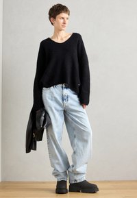 Suéter negro de punto esponjoso con mangas anchas, combinado con jeans sueltos de color azul claro y botines negros gruesos. Sosteniendo una chaqueta negra.