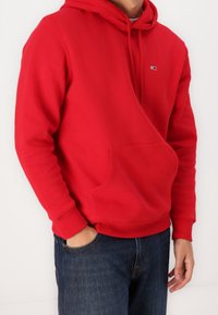 Sudadera roja de mezcla de algodón, con bolsillo canguro y puños acanalados, con un pequeño logo en el pecho. Combinada con vaqueros azules.