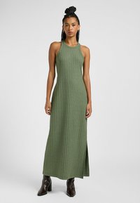 Abito maxi verde a coste con scollatura alta e spacco laterale. Presenta un corpetto aderente e scende fino alle caviglie; abbinato a stivali con tacco scuro.