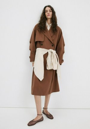 Mujer con cabello largo y rizado, vestida con un abrigo vestido marrón oversize, una camisa beige a cuadros atada alrededor de la cintura y zapatillas marrones con pulsera en el tobillo.