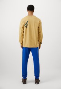 Beżowa koszula z długim rękawem z wyszywanym motywem ptaka na plecach, połączona z niebieskimi spodniami z bocznymi paskami i brązowymi butami o fakturowanym wzorze.