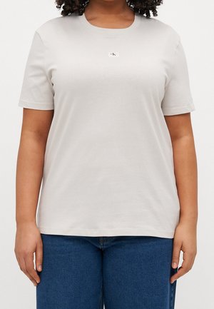 Person iført en ensfarvet lys grå t-shirt med korte ærmer med en lille Calvin Klein-logo nær halsudskæringen og blå jeans.