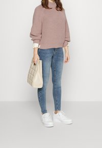 Pull en tricot rose clair à manches courtes, associé à un jean skinny bleu et des baskets blanches. Tenant un sac à main beige texturé.