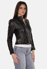 Giubbotto da moto in pelle nera con strisce verticali, chiusura zip, design aderente e dettagli in rete trasparente sulle maniche. Indossato sopra un top bianco.