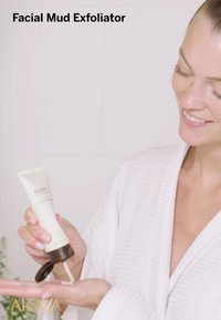 Ansiktsmud exfolierare i en vit tub med ett brunt lock, utdelad i en hand. Modell i vit badrock, leende, som visar upp texturen.