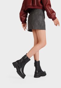 Stivali alla caviglia in pelle nera con suola spessa, chiusura con lacci e dettagli hardware a fessura. Finitura liscia, design robusto e suola con grip testurizzato.