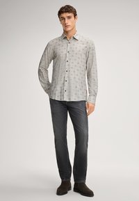 Jongeman in een lichtgrijs gestippeld overhemden met knopen, donkere grijze jeans en donkerbruine suède laarzen, staand voor een effen achtergrond.