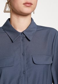 Femme portant une chemise bleu foncé boutonnée avec des poches poitrine et de grandes boucles d'oreilles triangulaires en chaîne dorée, fond neutre.