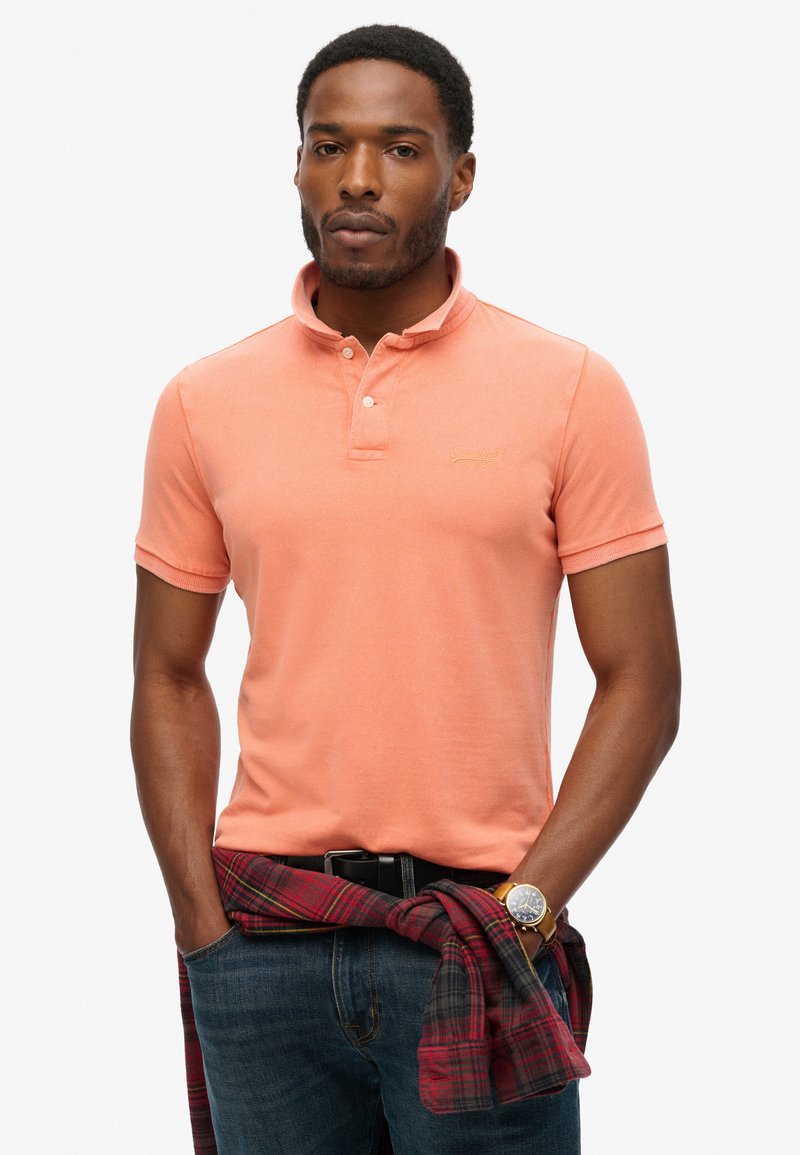 Licht koraalkleurig polohemd met een korte kraag, knopen en geborduurd logo. Gecombineerd met donkere jeans en een rood geruit overhemd om de taille geknoopt.