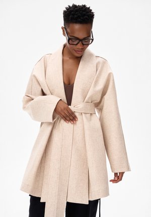 Person mit kurzen lockigen Haaren und Brille, trägt einen beigen Mantel mit Gürtel über einem braunen Oberteil und justiert den Mantelgürtel.