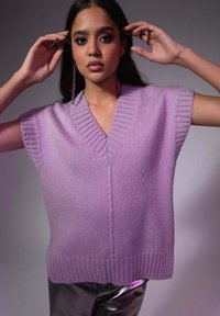 Gilet sans manches en tricot lilas avec un col en V, présentant des bords côtelés et une coupe décontractée. Le modèle pose les mains levées près du visage.