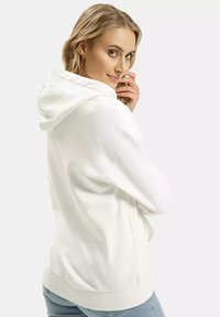 Biała bluza z kapturem o luźnym kroju, z teksturowanej tkaniny, z przednią kieszenią i ściągaczem u dołu. Nosi się ją na jasnoniebieskich dżinsach.