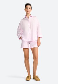 Light pink button-up shirt with rolled sleeves paired with light pink shorts. Model wears brown lace-up shoes. Simple and relaxed fit.