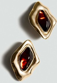 Zlaté náušnice s vlnitým okrajom a centrálnym tmavočerveným živicovým kameňom, so hladkými textúrami a asymetrickými tvarmi.