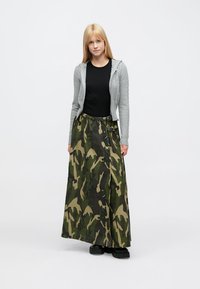 Falda larga camuflaje en tejido verde y beige con acabado texturizado. Combinada con un top negro y un cárdigan gris corto. Calzado negro.