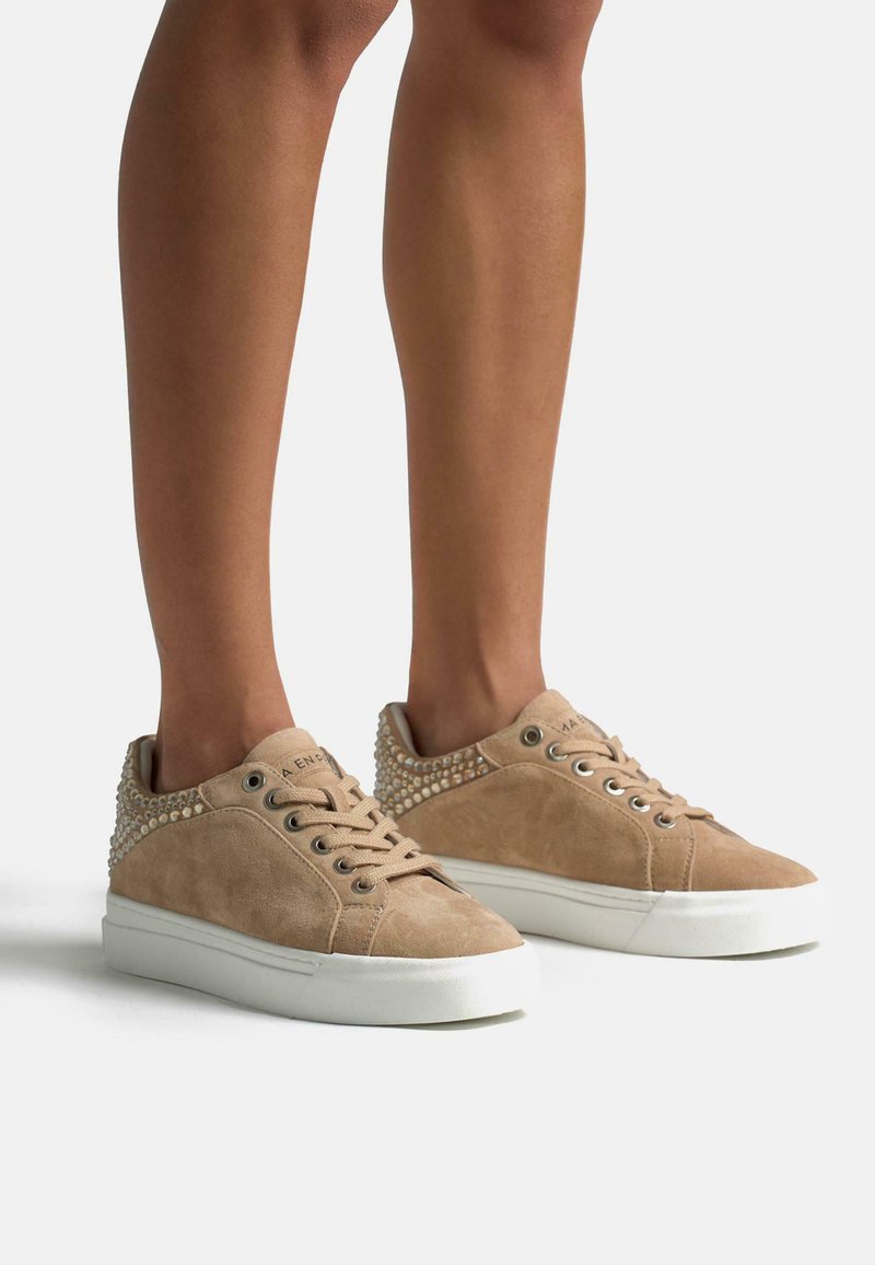 Tan suède sneakers met een gestructureerd oppervlak, ronde neus en decoratieve studs aan de achterkant, geplaatst op een witte rubberen zool.