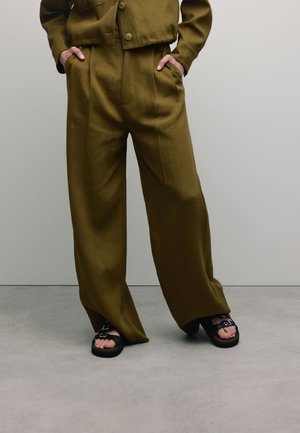 Olijfgroene, loszittende broek met plooien, gecombineerd met zwarte sandalen met een gesp-detail, getoond tegen een neutrale achtergrond.