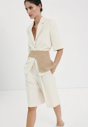 Vrouw draagt een crèmekleurige blazer met krijtstreepjes en ellebooglange mouwen, gecombineerd met bijpassende knielange shorts, afgewerkt met een brede, tan suède riem.