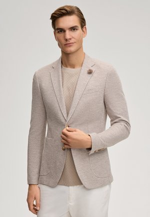 Hellbeiger Blazer aus strukturiertem Stoff, einreihiges Design, zwei Vordertaschen und dekorative Knopfakzente. Trägt man über einem beigen Hemd.
