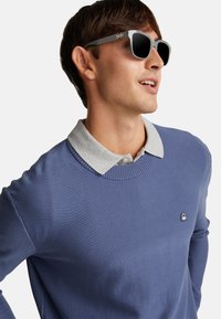 Jasnoniebieski sweter z prążkowanym dekoltem noszony na szarej koszuli z kołnierzykiem, zestawiony z szarymi okularami przeciwsłonecznymi z ciemnymi soczewkami i detalem emblematu z boku.