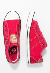 Baskets à enfiler en toile rouge avec bouts en caoutchouc blanc, œillets en métal, logos en patch de cuir marron, et semelles blanches à rayures noires sur fond blanc.