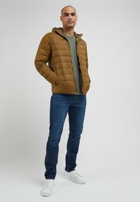 Męska brązowa kurtka puchowa z poziomym przeszyciem, szara podszewka, w zestawie z zieloną koszulką, ciemnymi dżinsami i bia�łymi trampkami.