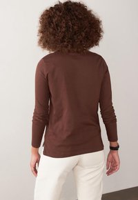 Brown long-sleeve top made of soft material, featuring a round neckline, fitted sleeves, and side slits. Worn with light-colored pants.