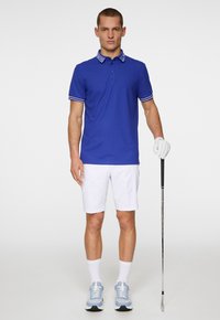 Royal blue polo shirt with striped collar, white shorts, and light blue sneakers. Golf club in right hand, wearing a white glove.