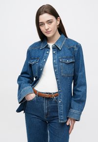 Μπουφάν τζιν με δύο τσέπες στο στήθος, χρώμα ανοιχτό μπλε, κλείσιμο με κουμπιά και παραδοσιακό γιακά· φοριέται πάνω από λευκή μπλούζα με μπλε τζιν.