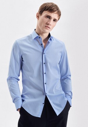 Chemise à manches longues bleu clair en coupe slim avec un col pointu, des boutons noirs et une texture lisse. Portée avec un pantalon sombre.