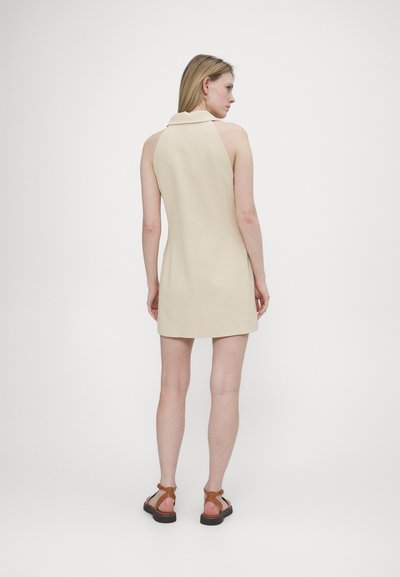 Beige ärmelloses Kleid mit Kragen, taillierter Passform und glatter Textur. Model trägt braune Riemchensandalen und wendet sich von der Kamera ab.