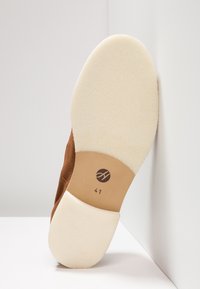 Semelle de chaussure en daim marron avec un dessous texturé de couleur crème. Présente un logo circulaire et la taille "41" imprim ée sur une surface lisse beige.