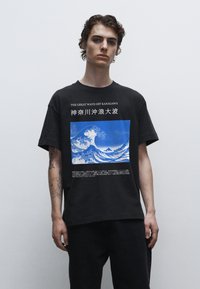 Černé bavlněné tričko s potiskem "Velká vlna u Kanagawy" v modré a bílé barvě, s anglickým a japonským textem pod obrazem.