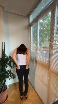 Femme aux longs cheveux foncés portant un débardeur blanc et un pantalon noir, debout les mains dans les poches arrière près des stores et des plantes d'intérieur.