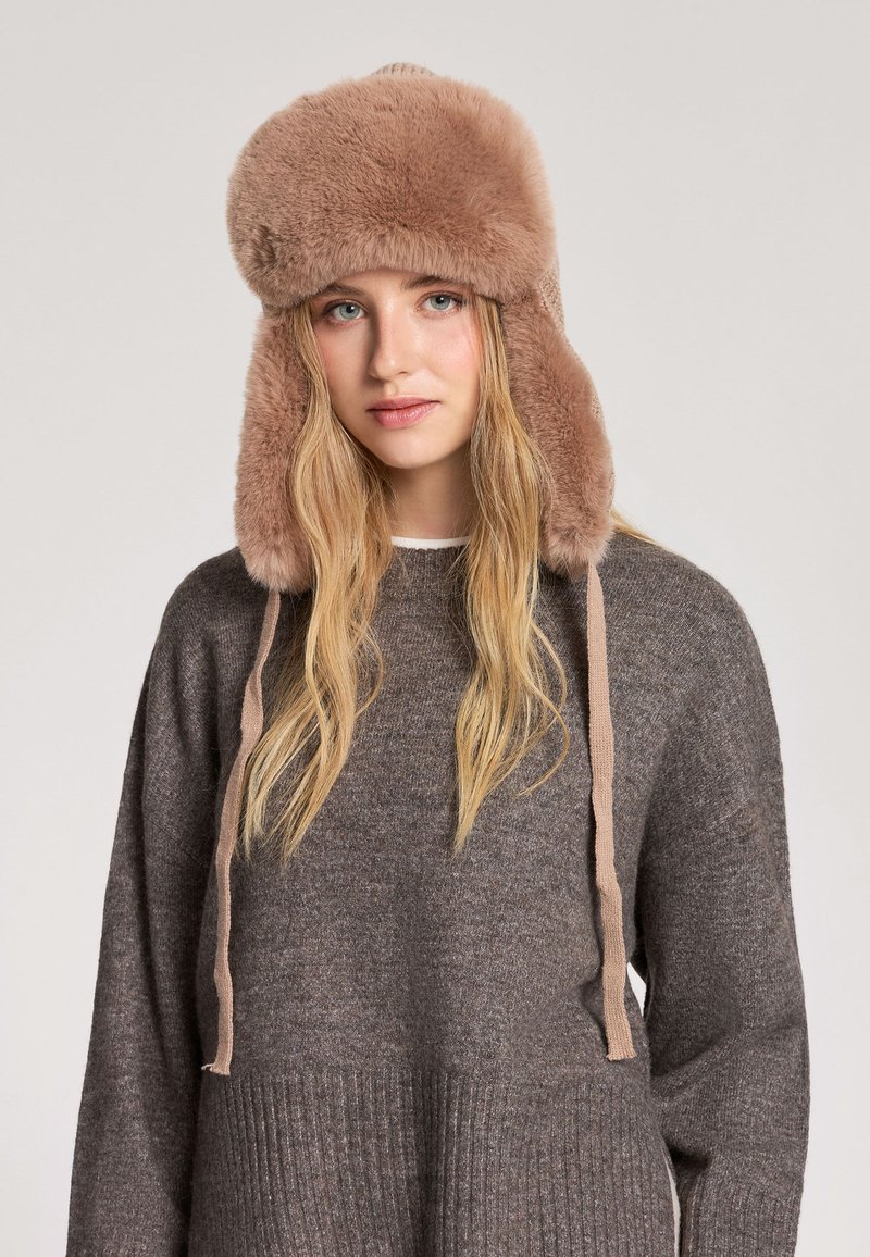 Brązowy futrzany kapelusz z nausznikami i trokami, o miękkiej fakturze i zaokrąglonej górze. Nosi się go z ciemnoszarym swetrem.