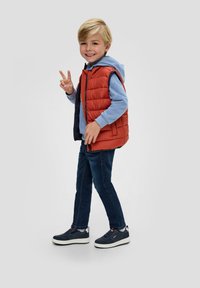 Niño con un chaleco acolchado rojo sobre una sudadera ligera azul, jeans oscuros y zapatillas marinas. El chaleco presenta un diseño acolchado y cremallera frontal.