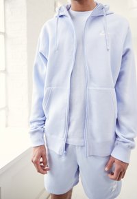 Ljusblå zip-hoodie med två framfickor och snörning, bärs över en vit T-shirt och matchas med ljusblå shorts.