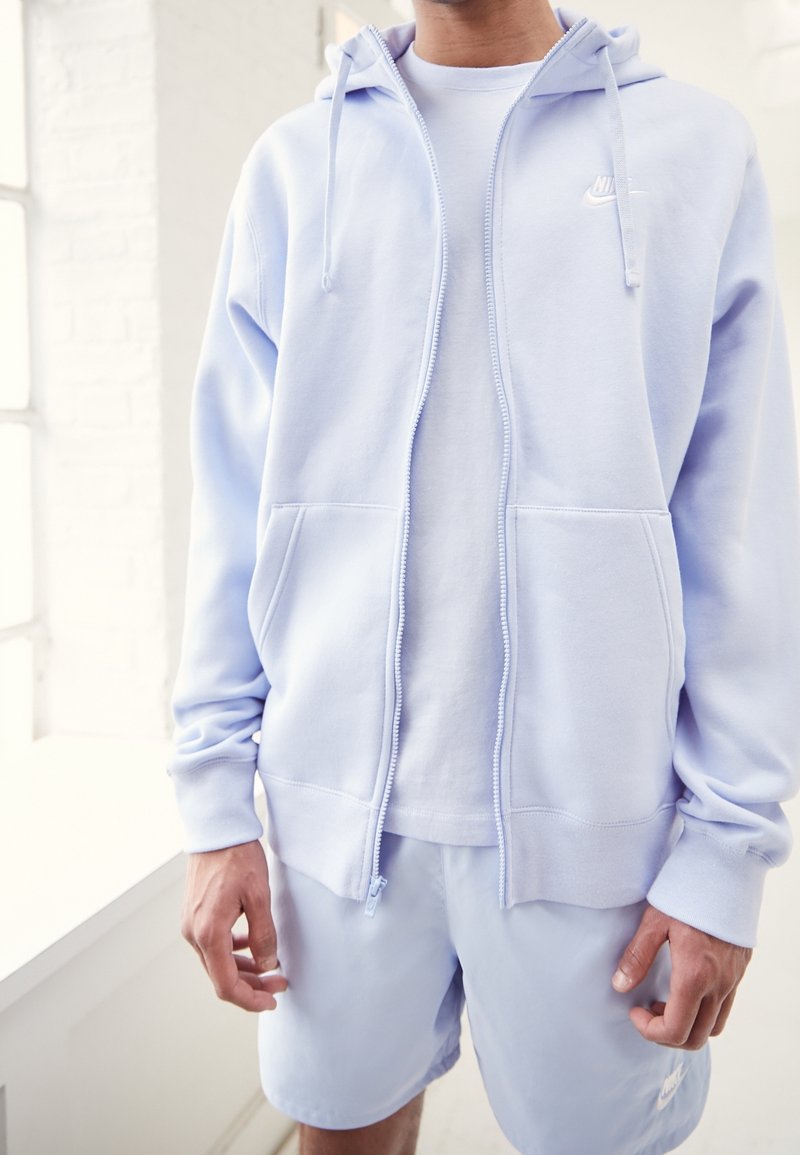 Ljusblå zip-hoodie med två framfickor och snörning, bärs över en vit T-shirt och matchas med ljusblå shorts.
