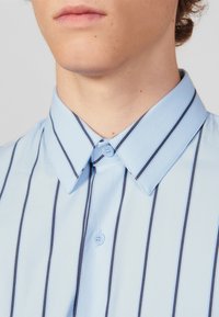 Camicia azzurro chiaro con righe verticali blu navy, colletto a punta e un solo bottone al collo. Tessuto liscio e vestibilità sartoriale.