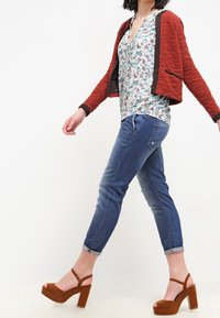 Cardigan rouge texturé court porté sur un chemisier à fleurs, associé à un jean slim bleu et des sandales compensées marron.