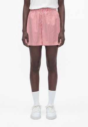 Person, die rote und weiße vertikal gestreifte Shorts, weiße Socken, weiße Turnschuhe und ein weißes Oberteil vor einem einfarbigen Hintergrund trägt.