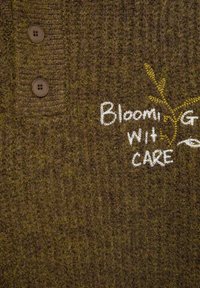 Braunes Strick-Polo mit einem strukturierten Muster, ausgestattet mit zwei braunen Knöpfen und gesticktem Text in Weiß und Gelb mit der Aufschrift "Blühend mit Sorgfalt."