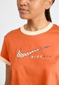 T-shirt en coton orange avec un col crème. Présente un logo Nike à carreaux et le texte "NIKE AIR ZOOM" en bleu en dessous.