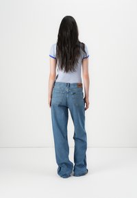 Persona con lunghi capelli scuri che indossa una maglietta a righe e jeans larghi, di spalle contro uno sfondo bianco semplice.