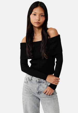 Młoda kobieta z długimi ciemnymi włosami, ubrana w czarny prążkowany sweter z odkrytymi ramionami oraz jasnoszare wysokiego stanu jeansy, pozująca na jednolitym tle.